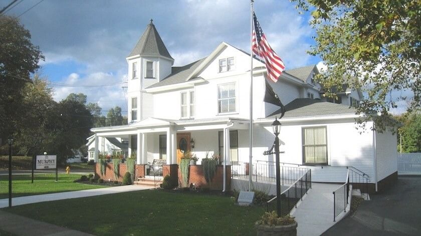 White building with porch, turret, American flag, ramp for accessibility, and a green lawn.