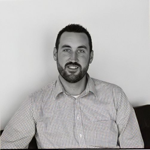 Man in a checkered shirt seated against a plain white wall, facing the camera and smiling gently