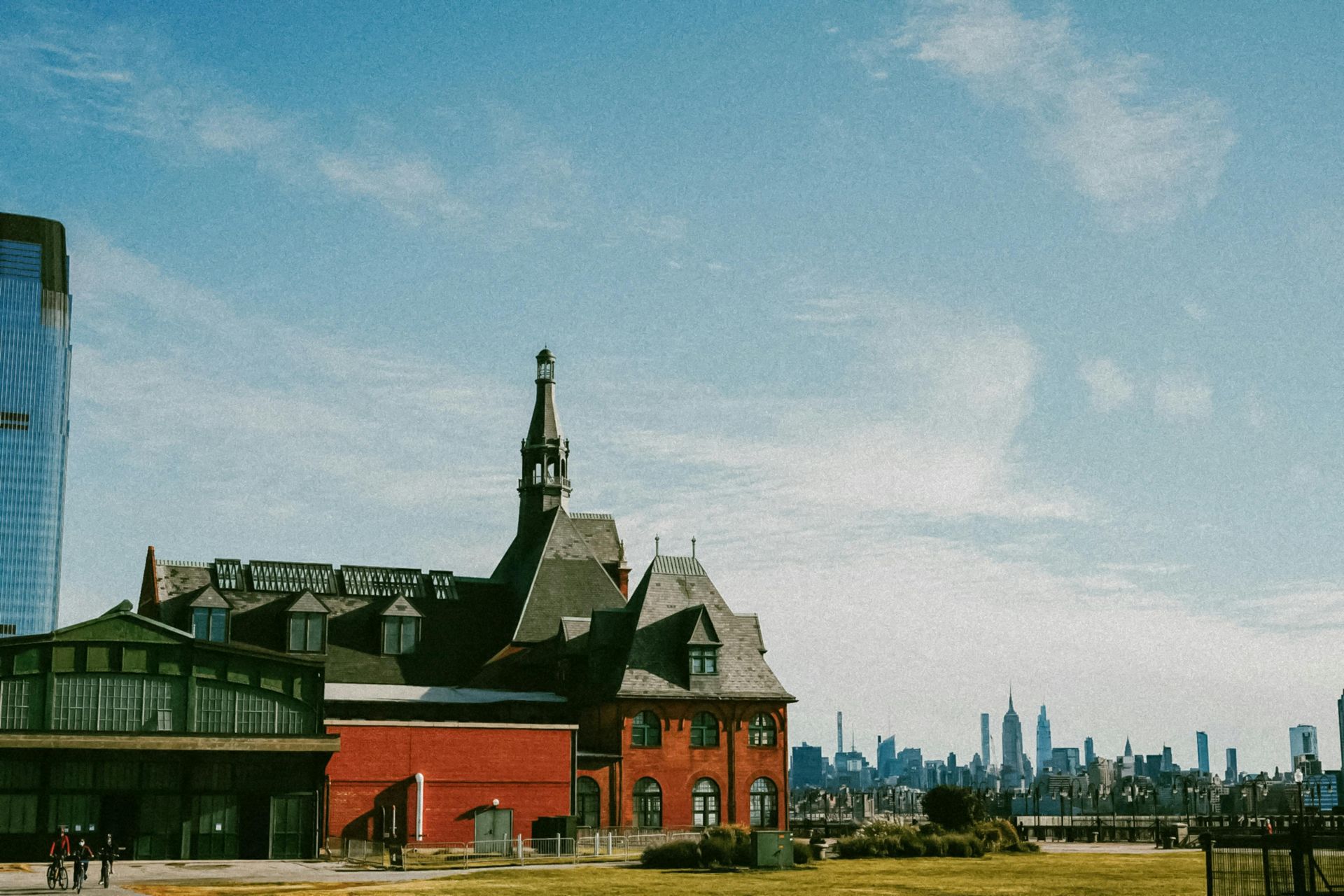 Historic red-brick building by water with a city skyline in the distance under a blue sky