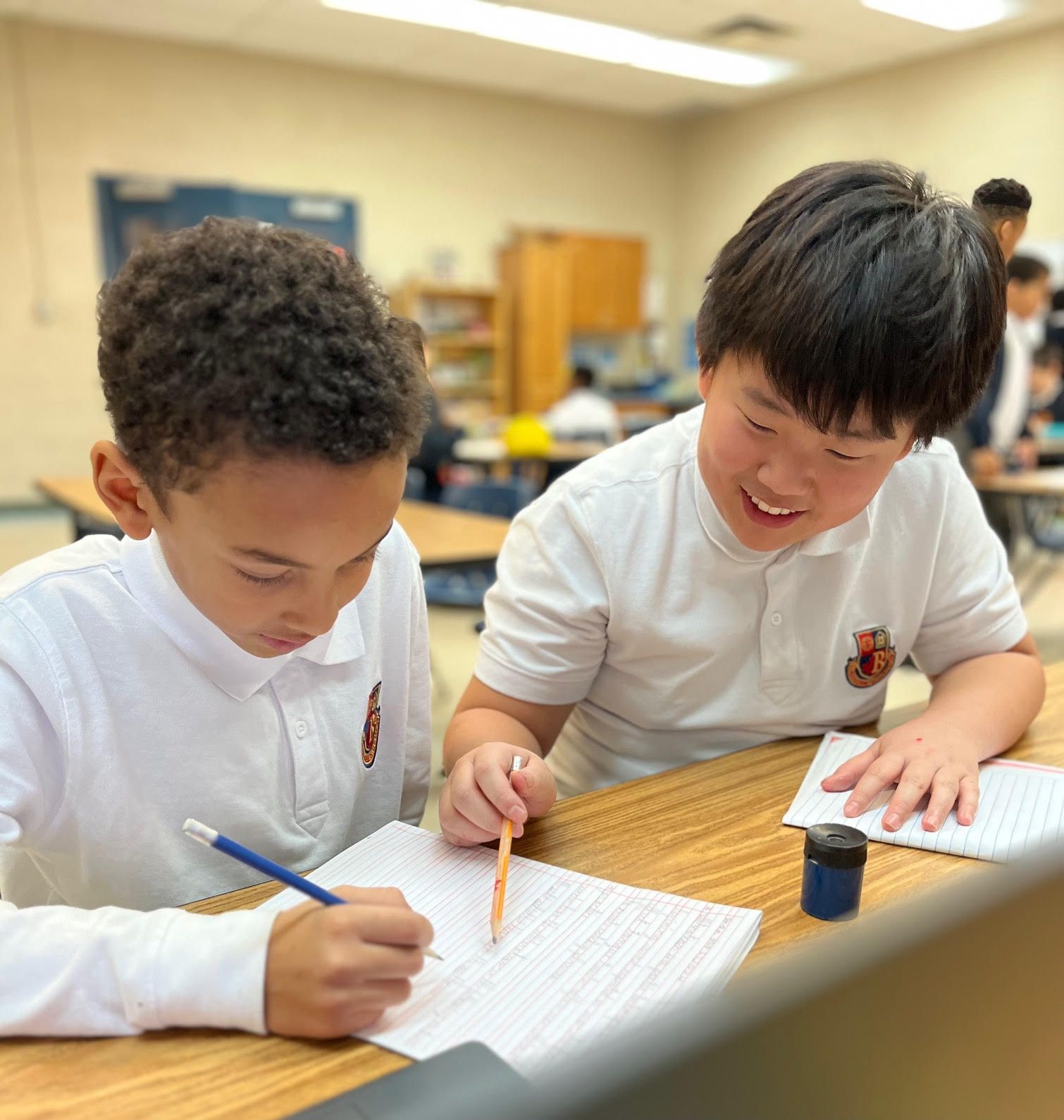 Two young boys are sitting at a desk writing in a notebook