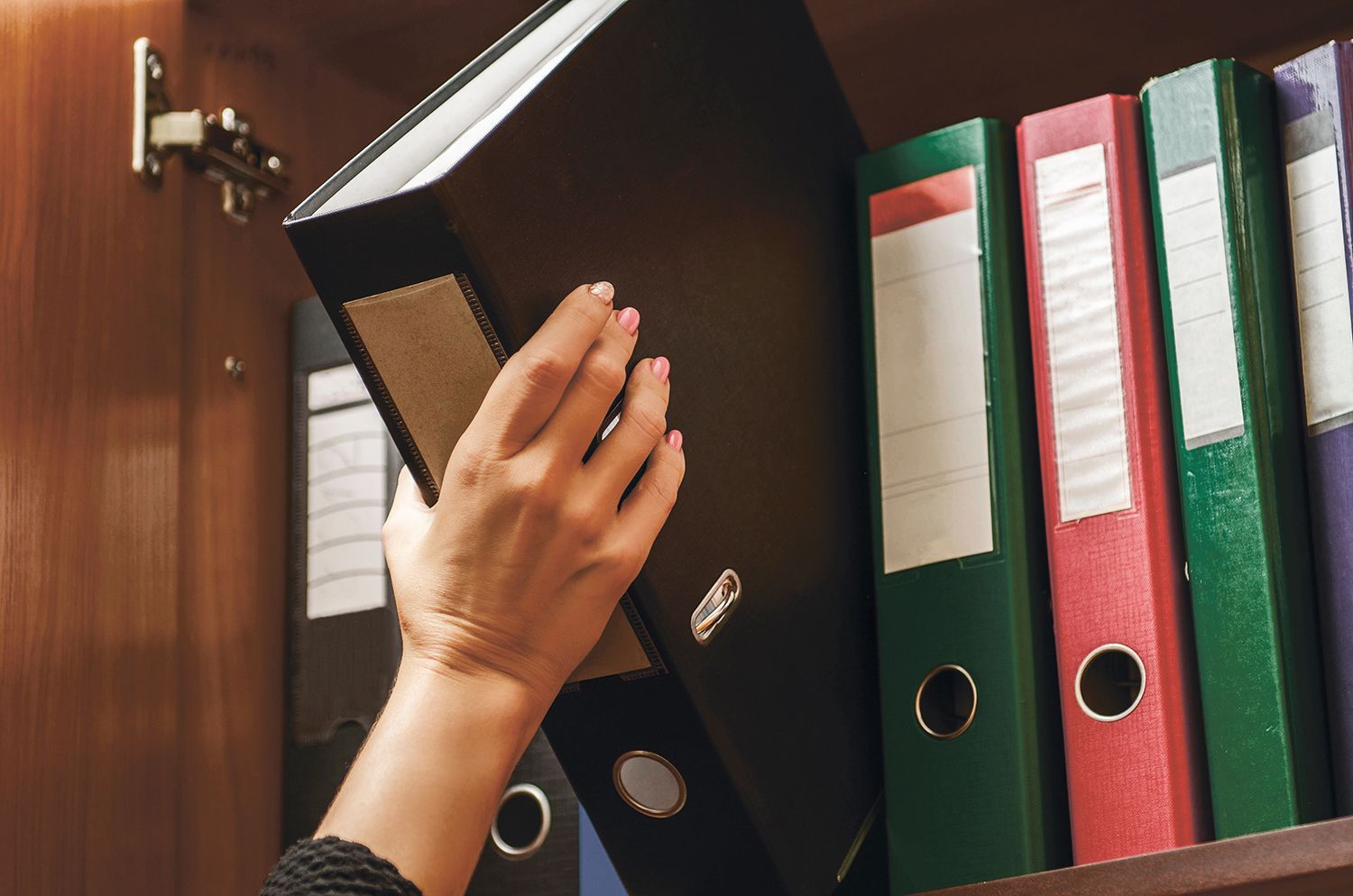 Hand placing a black binder on a shelf beside red and green ring binders