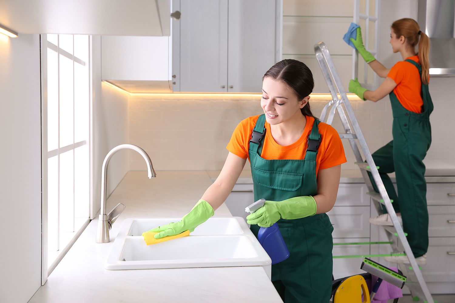 Two cleaners in orange shirts and green aprons cleaning a bright kitchen, one wiping a counter and one on a ladder.