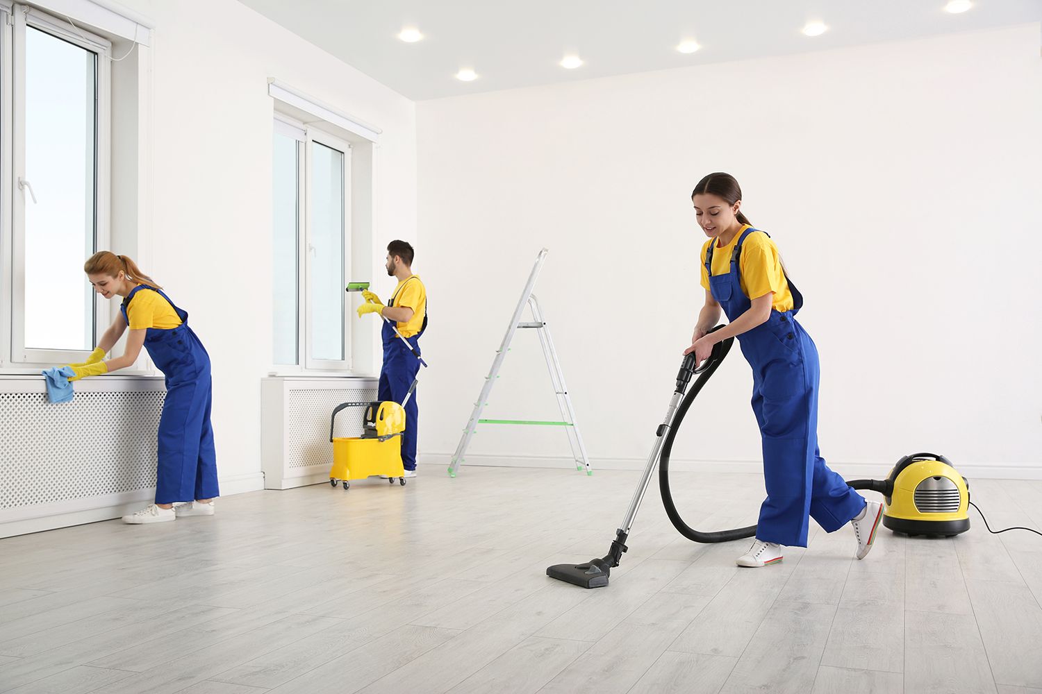 Three cleaners in blue uniforms vacuum and wash floors and windows in a bright white room.