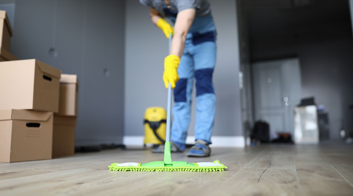 Person mopping a hardwood floor in a room with moving boxes and a yellow vacuum in the background