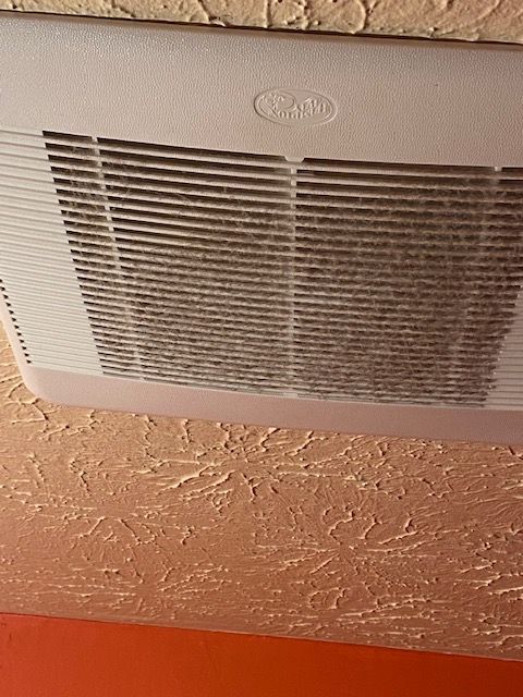 White ceiling vent grille on a textured peach wall with a small oval logo above it