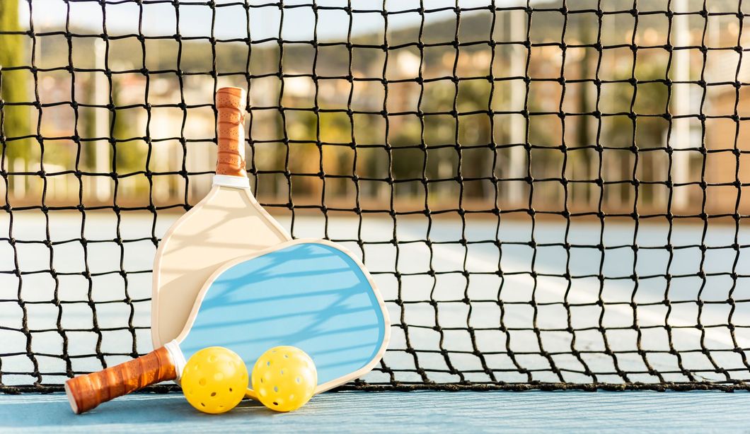 Tennis racket, ball, and two yellow balls leaning against a net on a court