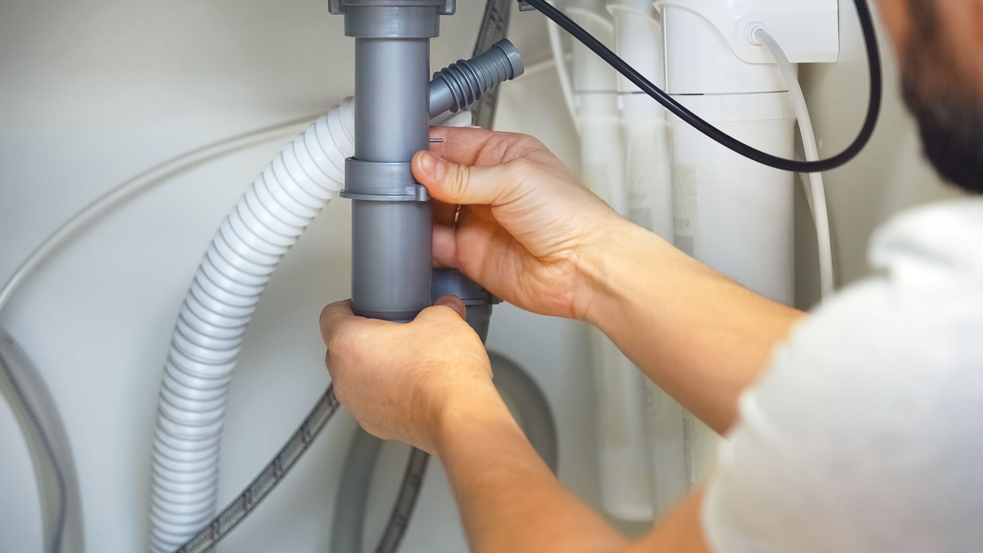 Hands adjusting a gray pipe under a sink with white hoses in a utility area