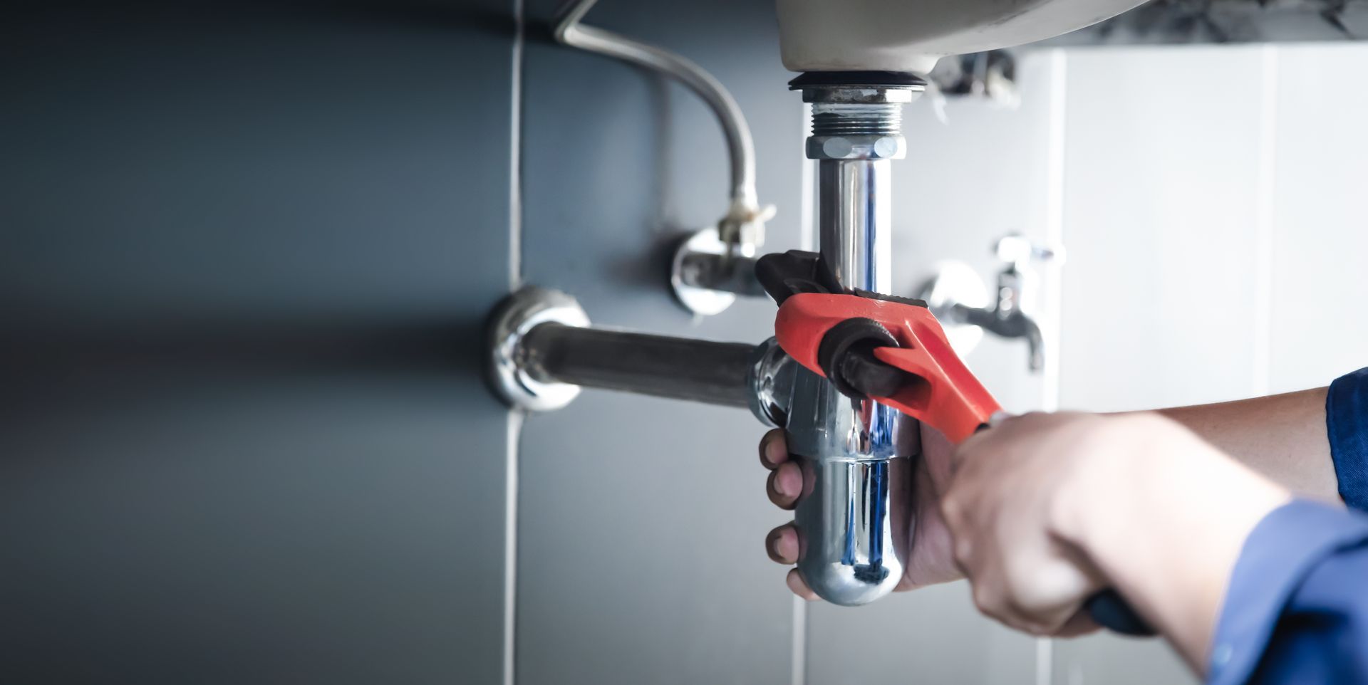 Hands use a wrench to tighten a chrome pipe connection under a sink.
