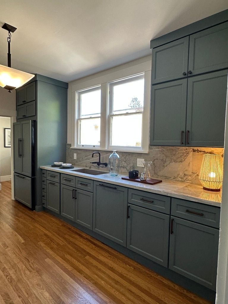 A galley-style kitchen featuring dark blue cabinets, marble countertops, light wood floors, and windows over the sink.