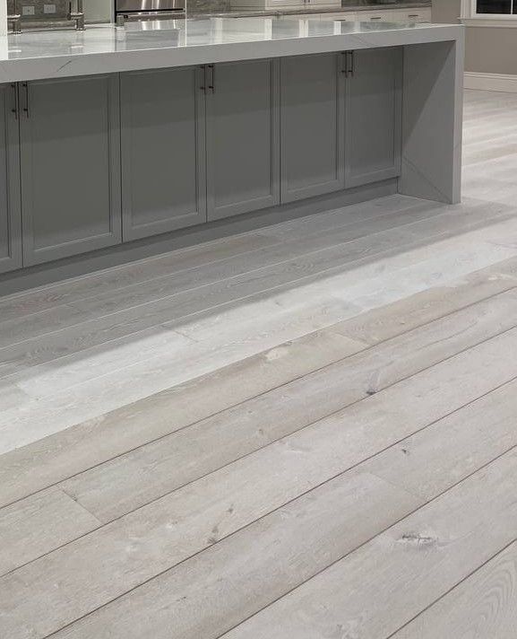A kitchen island with grey cabinets and a white countertop, positioned above light-toned, wide-plank wood flooring.