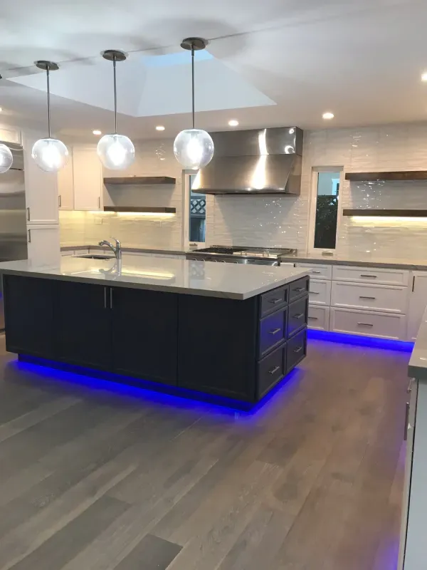 A modern kitchen featuring a dark island with blue LED lighting, white cabinetry, wooden shelves, and hanging globe lights.