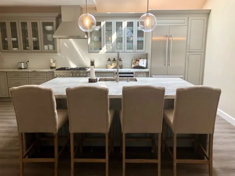A modern kitchen with a large marble-topped island, four beige upholstered bar stools, gray cabinetry, and a range hood.