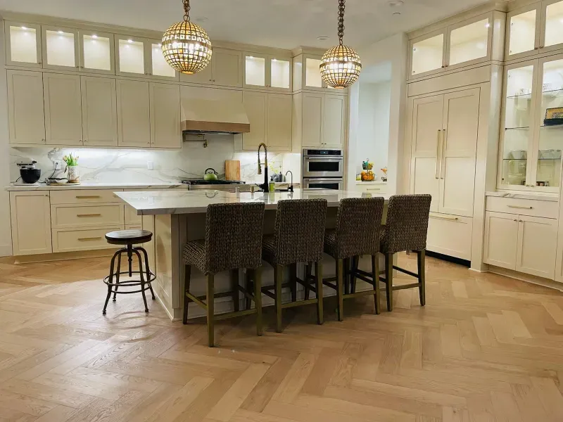 A spacious kitchen with white cabinetry, a center island with four dark stools, and light-toned herringbone wood floors.