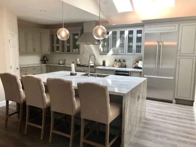 Modern kitchen with a marble-topped island, four beige bar stools, gray cabinetry, and stainless steel appliances.