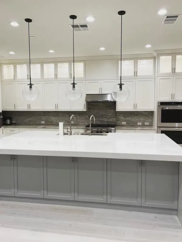 Modern kitchen featuring a large gray island with white countertops, white upper cabinets, and three hanging glass lights.
