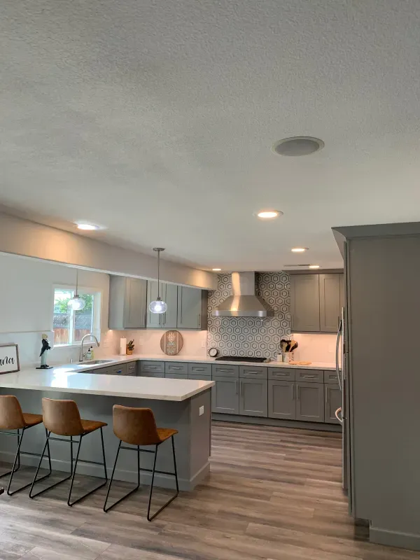 A modern kitchen with grey cabinets, quartz countertops, a tiled backsplash, and three brown bar stools at an island.