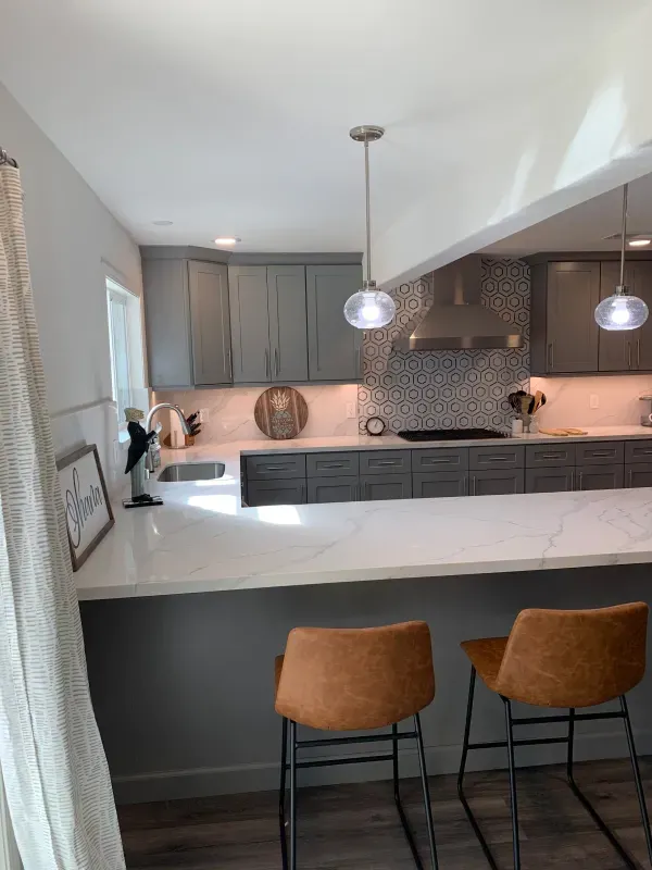 A modern kitchen with grey cabinets, white marble-style counters, a mosaic backsplash, and two tan bar stools.