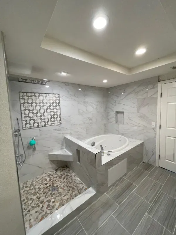 Modern bathroom with a glass-enclosed shower featuring a stone pebble floor, next to a white soaking tub and gray tile.