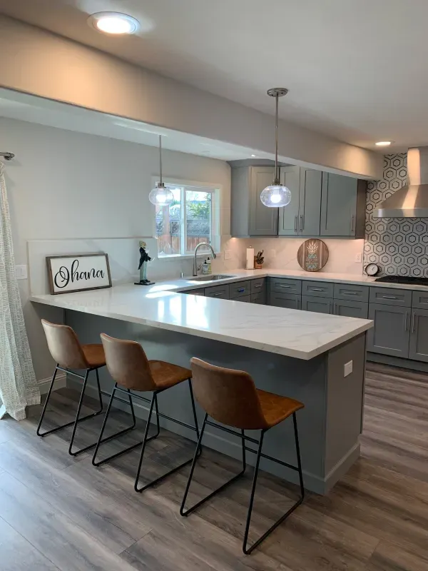 A modern kitchen with a grey U-shaped island, white countertops, three brown bar stools, and grey cabinetry.