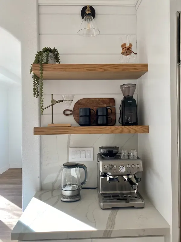 A home coffee bar with two wood floating shelves above a stone counter holding an espresso machine and a glass kettle.