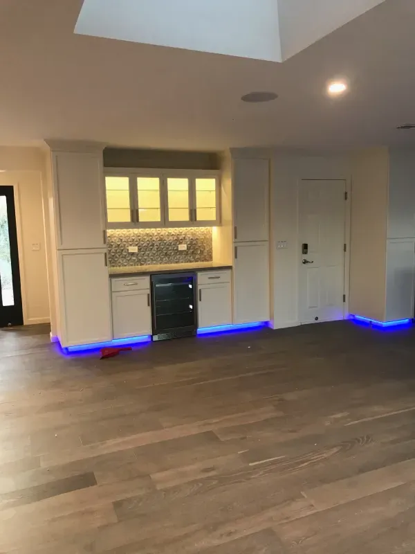 A modern home bar with white cabinets, glass-front upper storage, a wine fridge, and blue under-cabinet accent lighting.