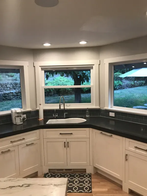 A kitchen corner featuring a white corner sink, black countertops, white cabinets, and large windows looking outside.