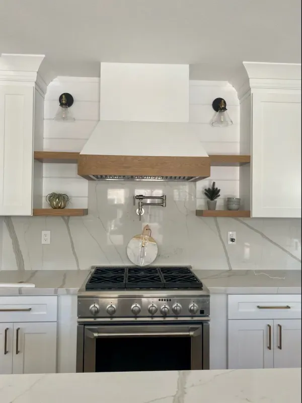 A modern kitchen stove area with white cabinets, a wood-trimmed range hood, open shelves, and marble-style backsplash.