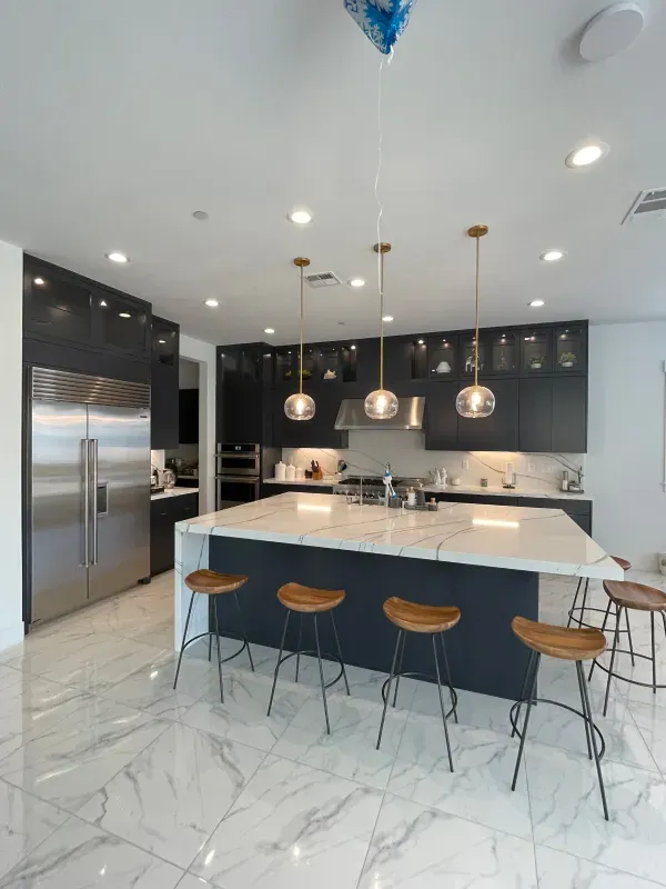 A modern kitchen featuring dark cabinetry, white marble-style flooring, a large marble island, and four wooden bar stools.