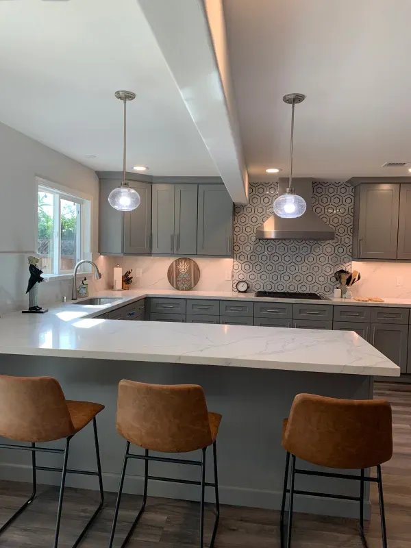 A modern kitchen with grey cabinets, white countertops, three brown bar stools, and two hanging glass pendant lights.