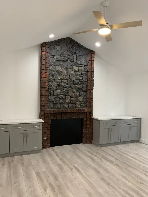 A stone fireplace with a red brick frame centered between grey storage cabinets in a room with light wood-look flooring.