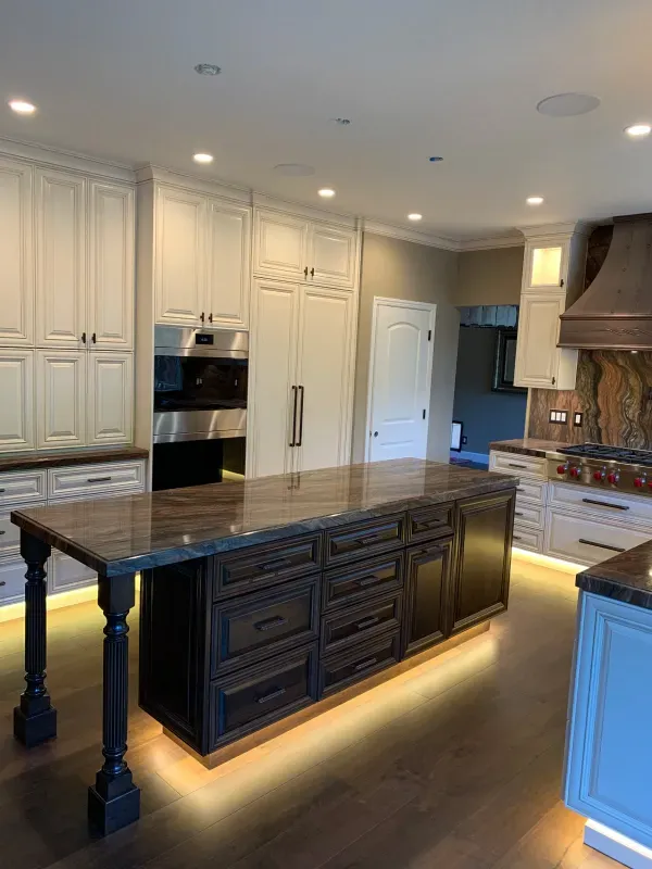 A modern kitchen featuring a dark wooden island with under-cabinet lighting and white cabinetry against beige walls.