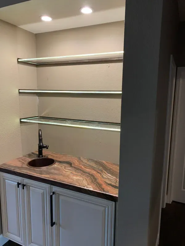 A wet bar with white cabinets, a brown stone countertop, a sink, and three illuminated floating glass shelves.