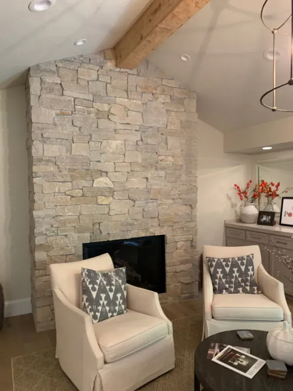 Two cream-colored armchairs with patterned cushions face a stone fireplace in a cozy, neutral-toned room with a wood beam.