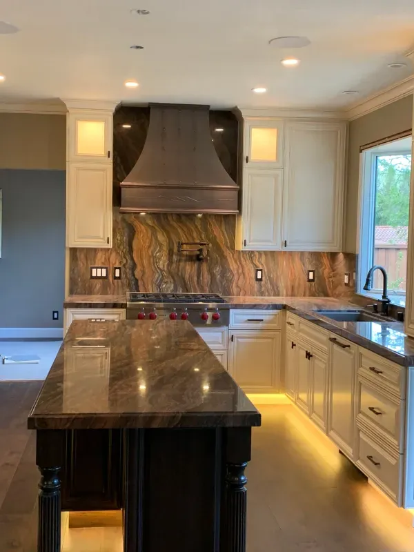 A modern kitchen with white cabinets, dark wood island, contrasting stone backsplash and countertops, and under-cabinet lighting.