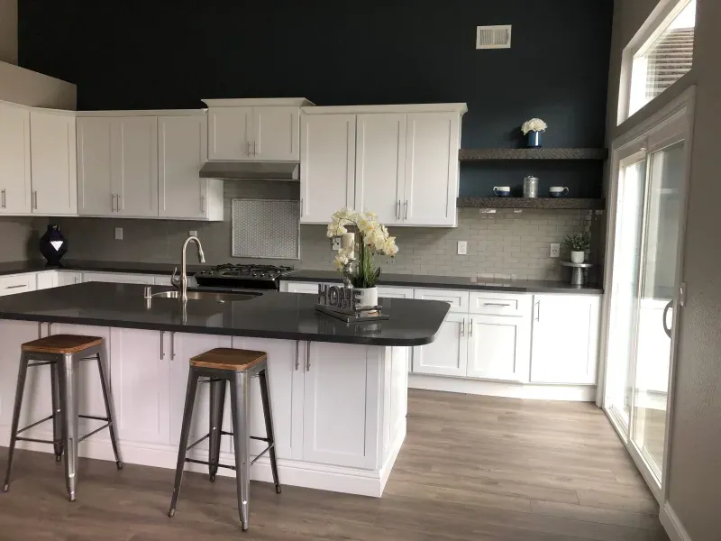 Modern kitchen with white cabinets, dark countertops, two metal bar stools at an island, and a dark accent wall.