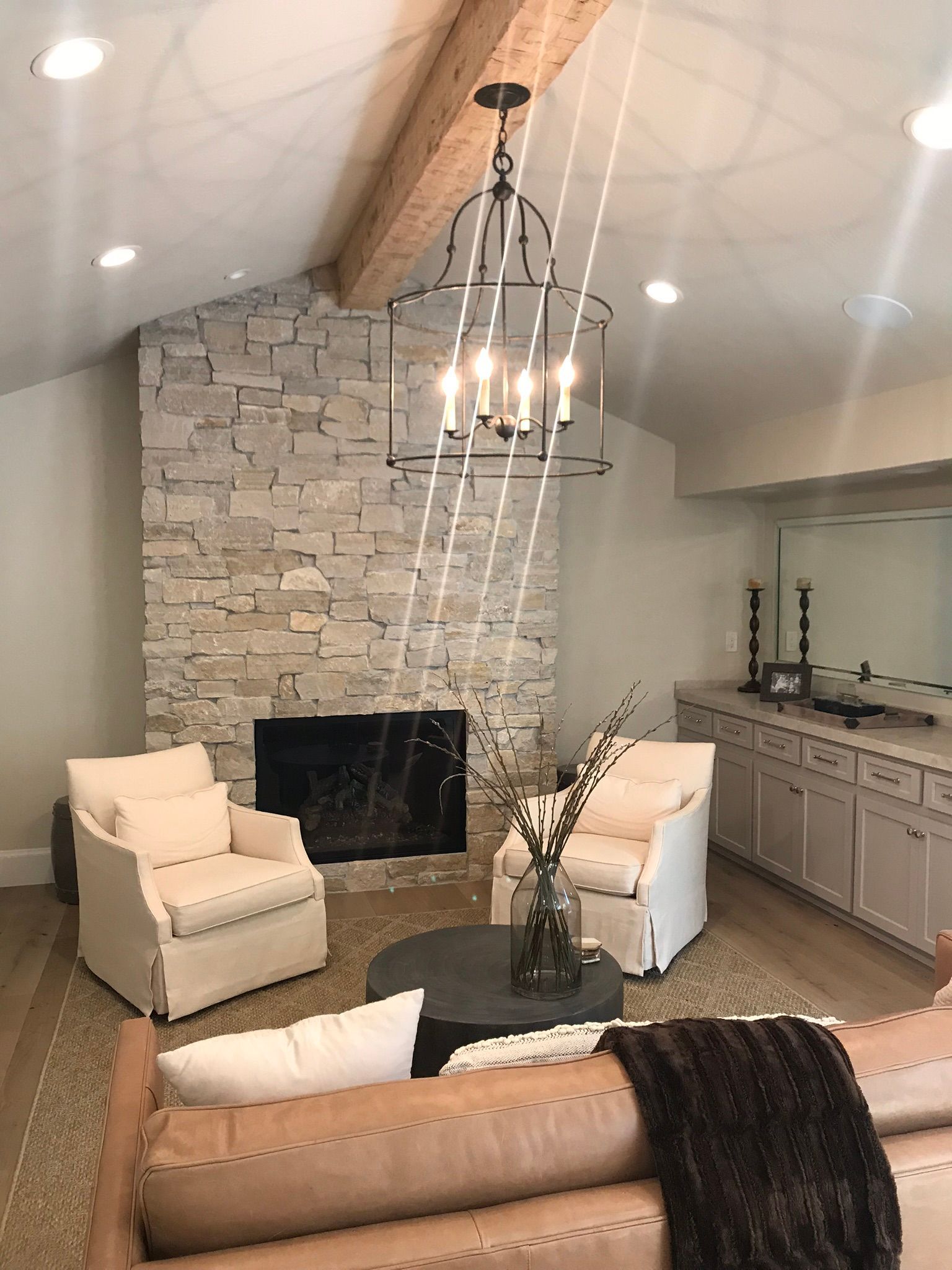 Living room with a stone fireplace, cream armchairs, a round coffee table, and a light brown sofa under a wood ceiling beam.