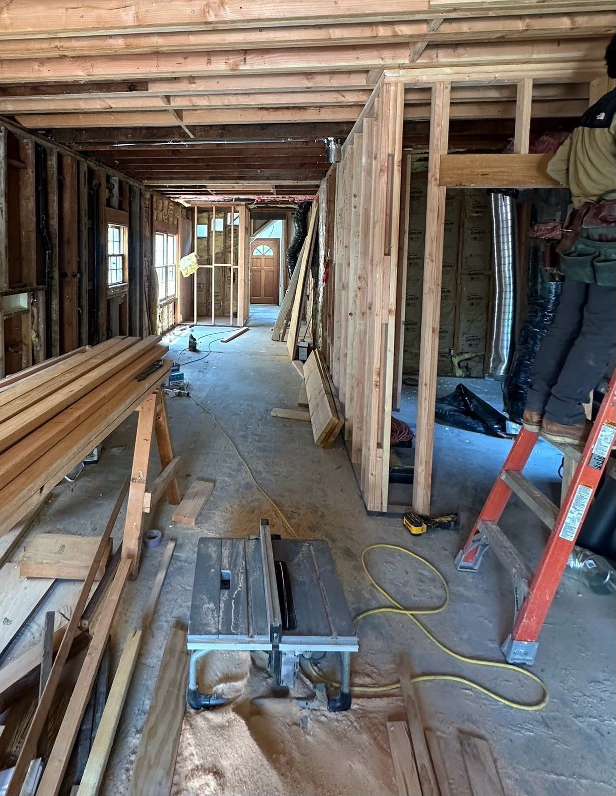 An interior renovation site with exposed wall studs, lumber piles, a table saw, and a person working on a ladder.