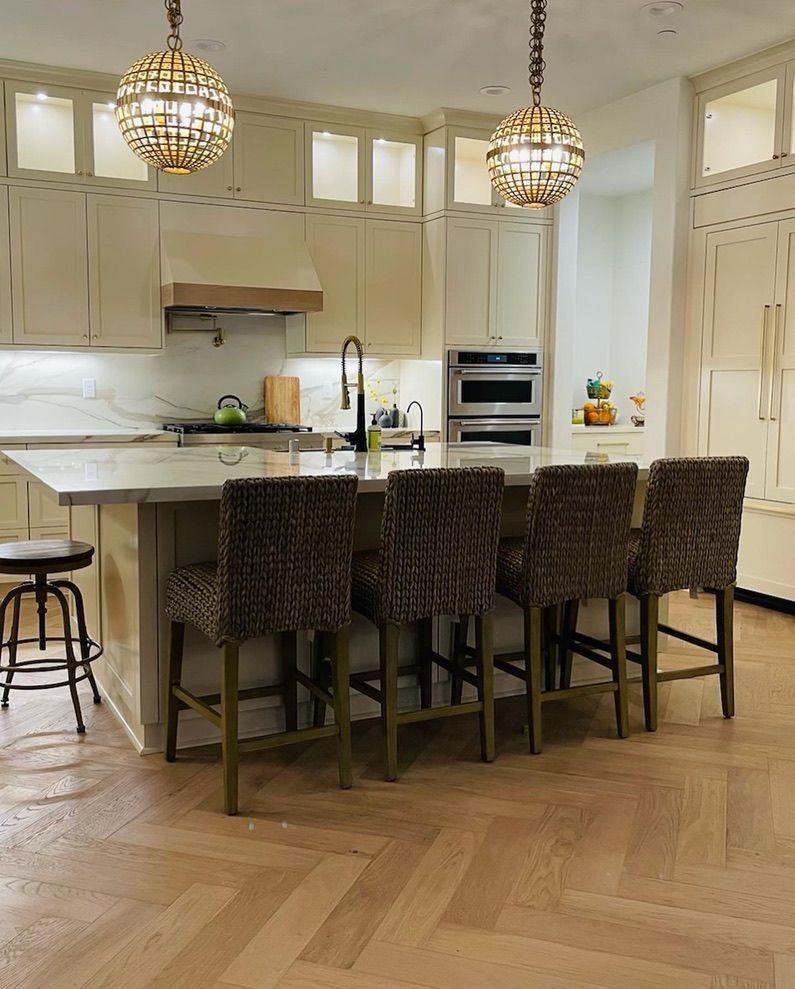 A kitchen with a large island, four dark patterned bar stools, light cabinetry, and two spherical chandeliers.