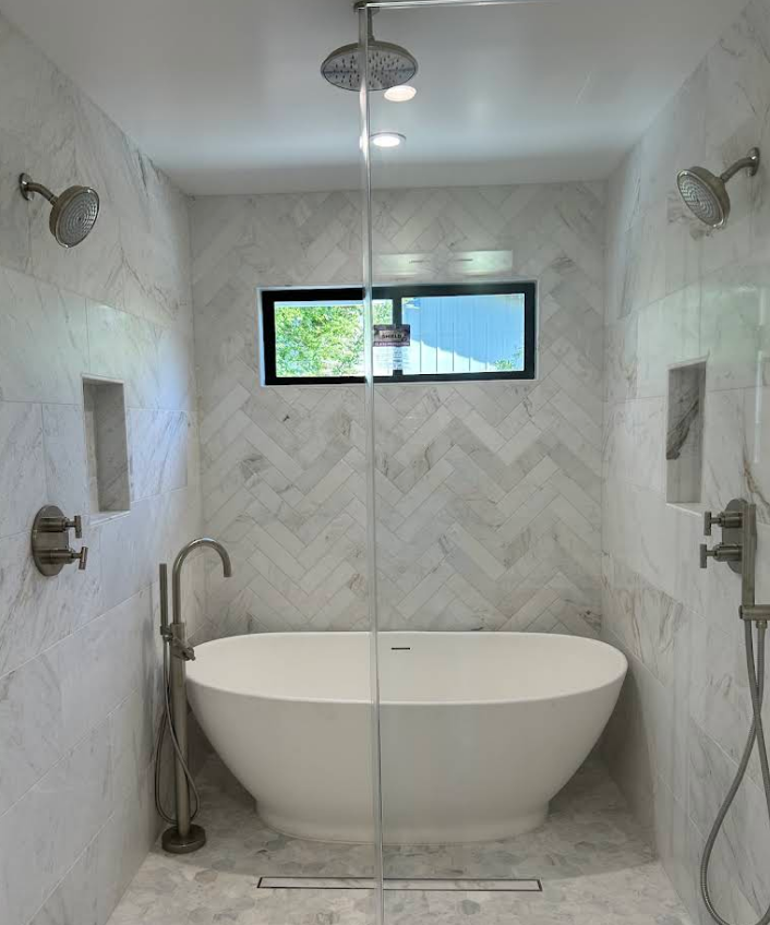 A modern walk-in shower with a freestanding white tub, marble tile walls in a herringbone pattern, and silver fixtures.
