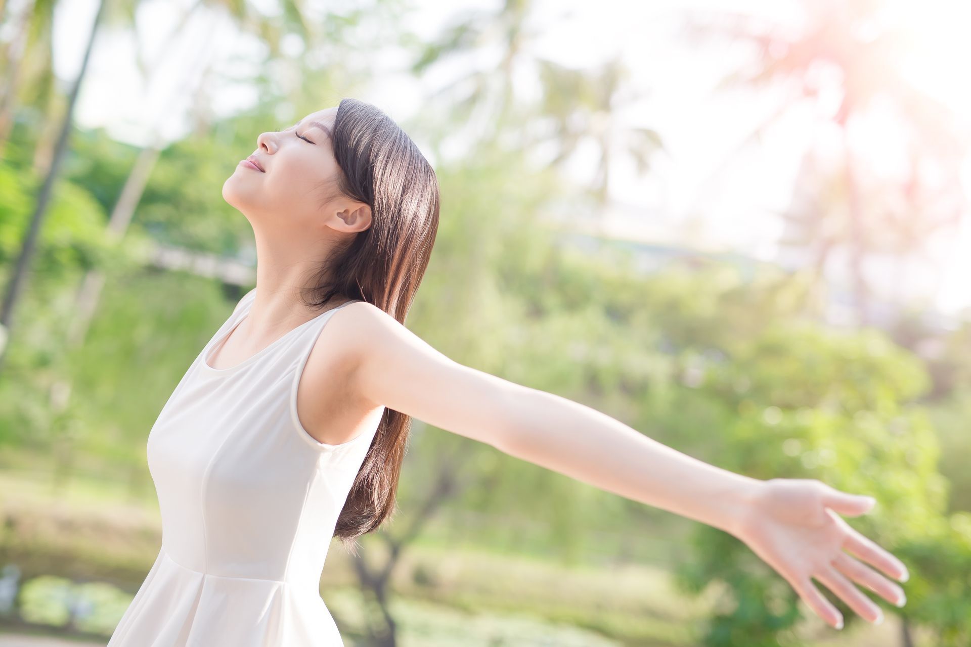 Woman with arms outstretched, eyes closed, enjoying sunlight in a green, outdoor setting.