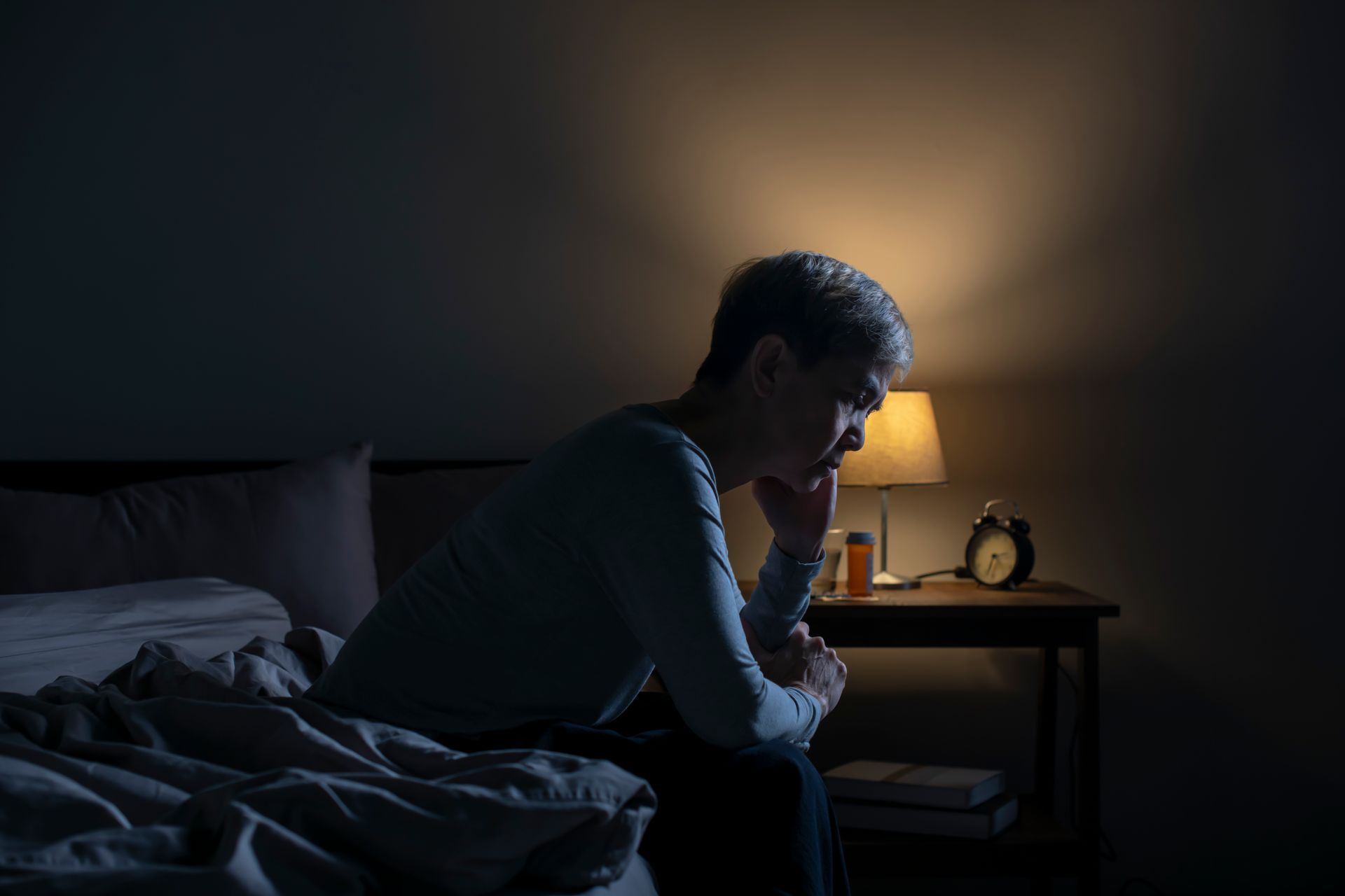 Person sitting on bed, looking down, in a dimly lit bedroom with nightstand and lamp.