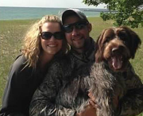 A couple posing for a picture with their dog in a scenic outdoor setting.