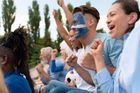 A group of excited sports fans cheering with arms raised at an outdoor stadium, one fan has face paint on.