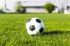 A black and white soccer ball sits on a vibrant green grass field with a blurred building in the background.