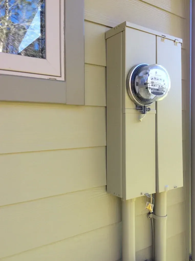 Electric meter box mounted on beige house siding beside a window