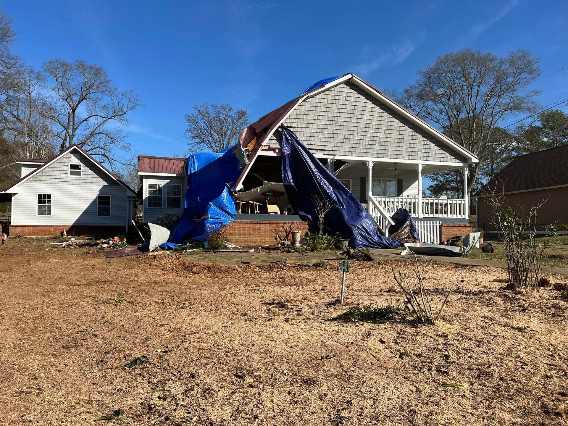 A house with significant roof damage covered by blue tarps, standing next to a smaller house under a clear blue sky.