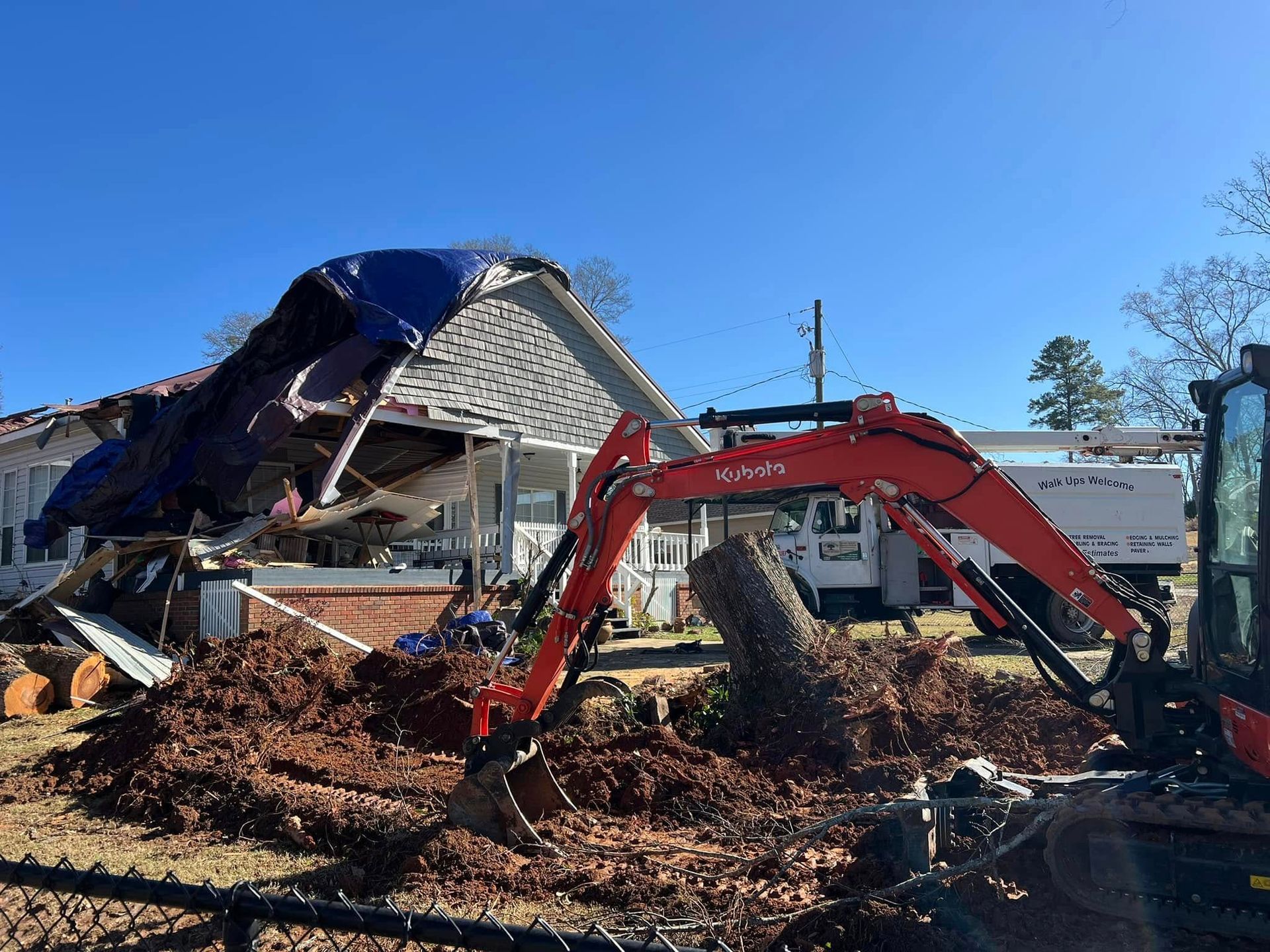 An orange excavator works in the yard of a house with a blue tarp over its damaged roof under a clear blue sky.