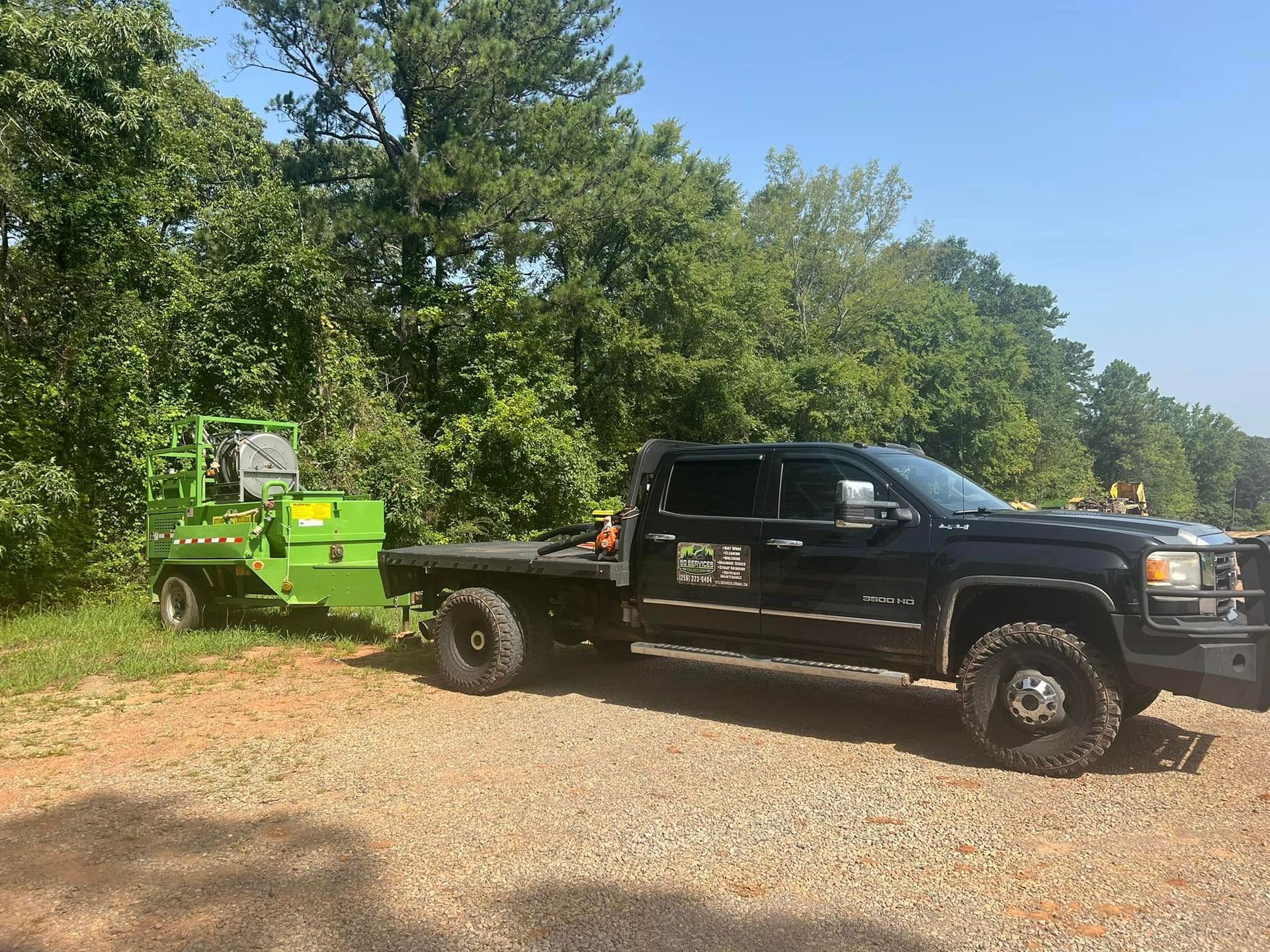 A black pickup truck parked on a gravel path pulling a bright green industrial wood chipper near a treeline.