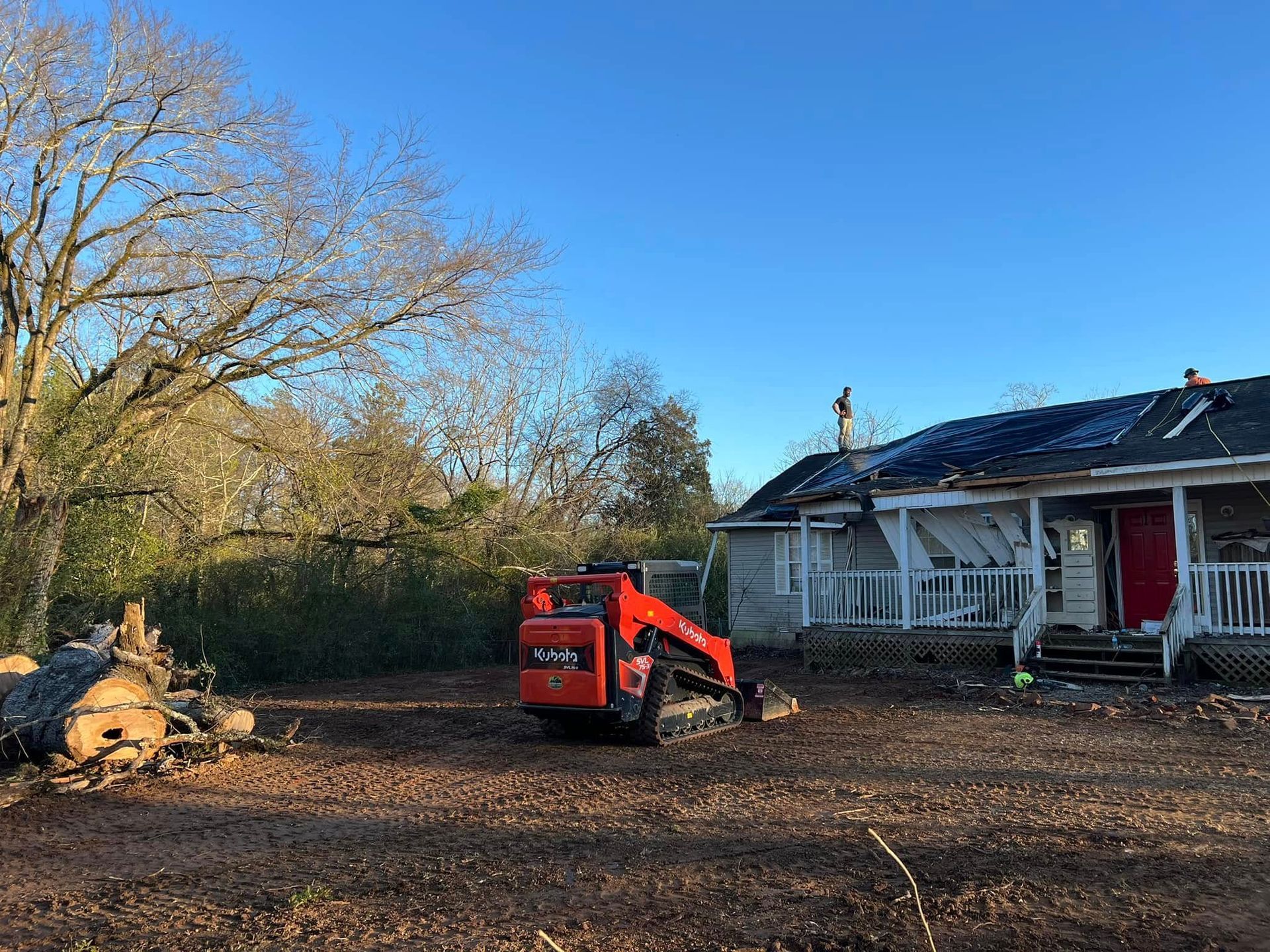 An orange skid steer loader sits on a dirt lot in front of a damaged, white-sided house under a clear blue sky.