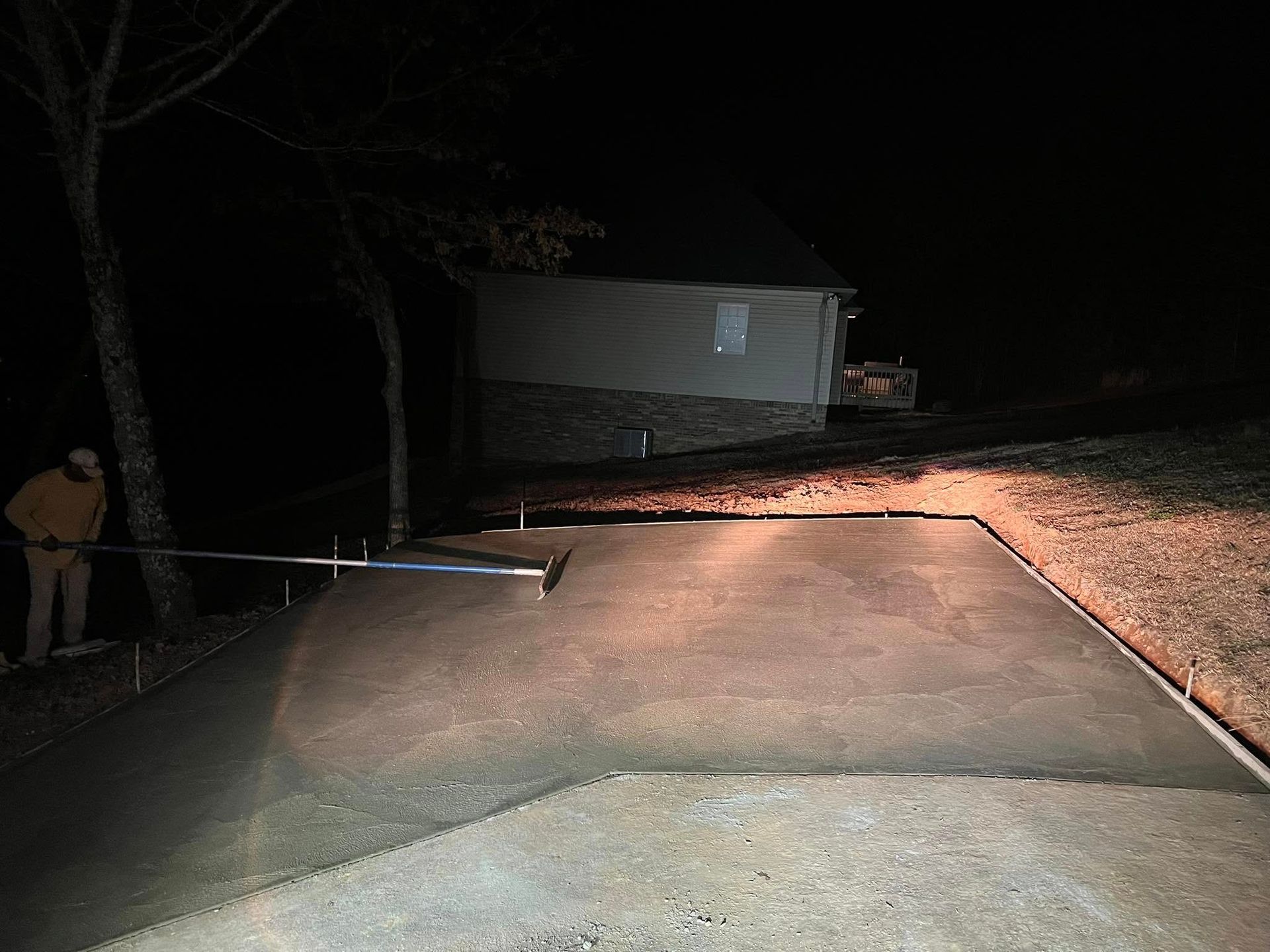 A person uses a tool to smooth freshly poured concrete on a residential driveway at night under artificial lighting.